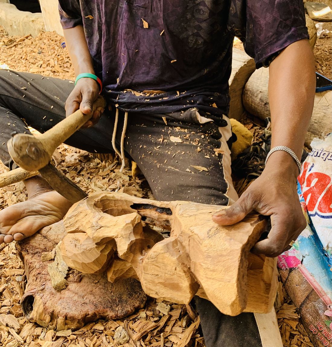 A woodcarver shaping a mask with an adze, surrounded by wood shavings